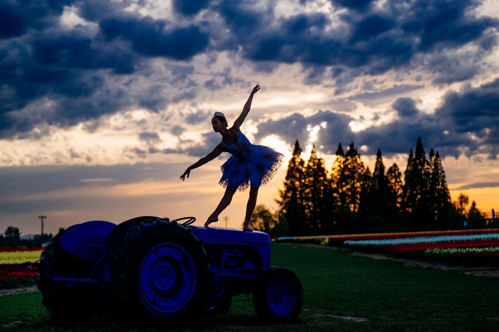 Female ballet dancer balances gracefully on a tractor in a rural field under a colorful sunset sky, USA.