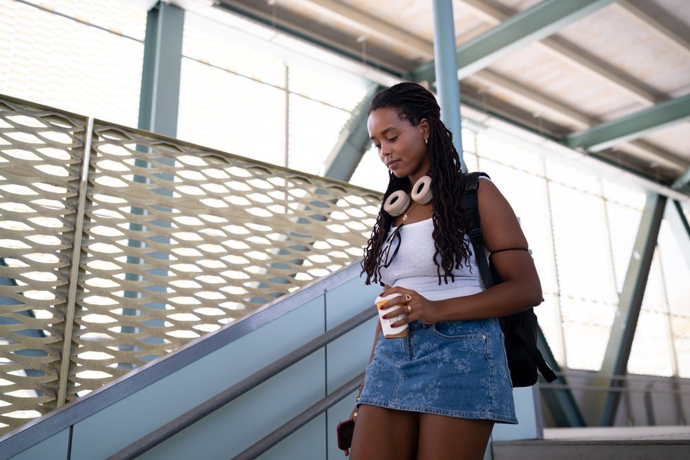 A young woman carrying a backpack and wearing headphones walks down outdoor stairs while holding a coffee cup, remote work, travel, Santiago De Compostela, Spain.
