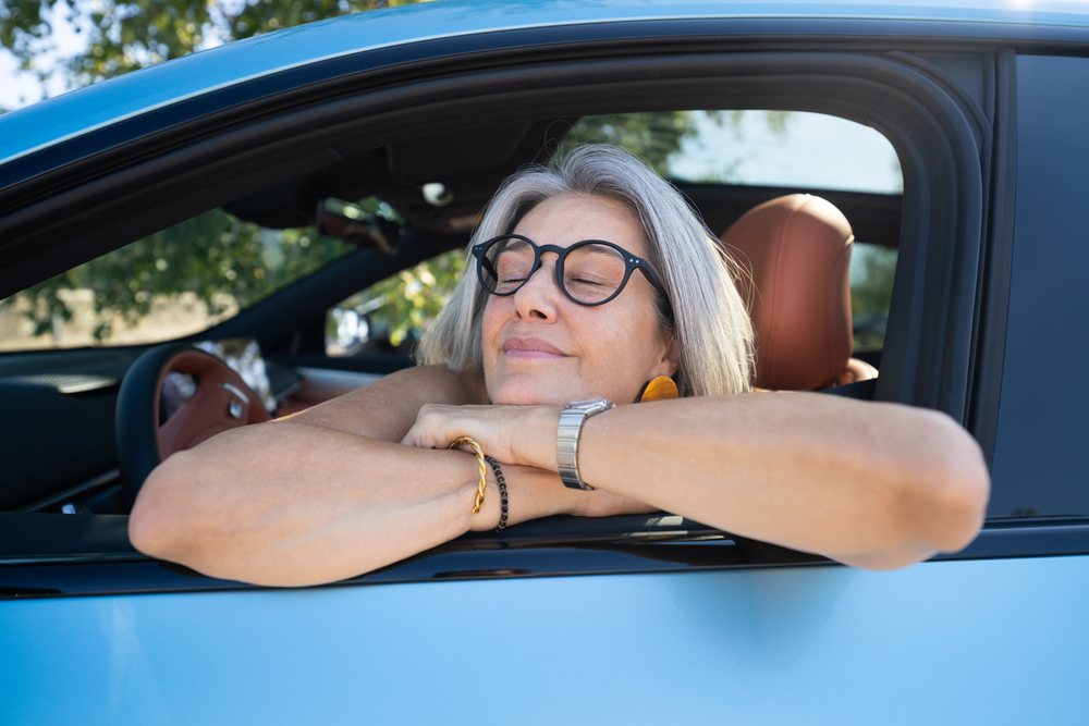 Mature woman with gray hair and glasses resting her arms on the window frame of a blue car outdoors during daytime, businesswomen, Spain.