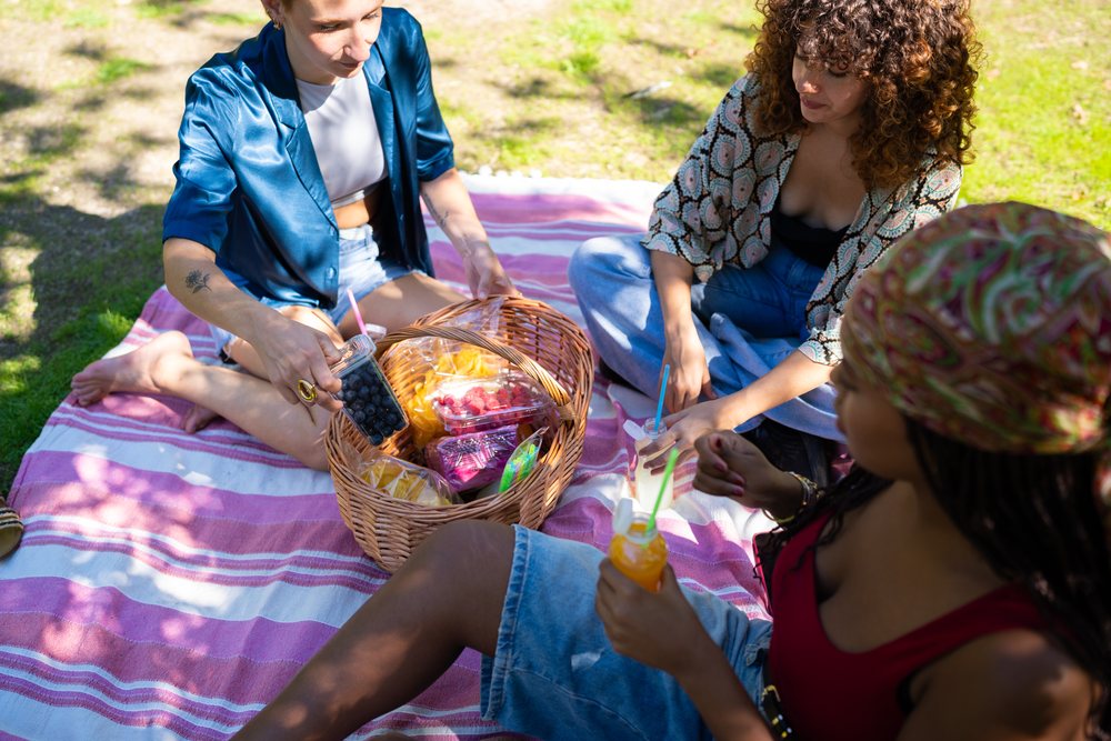 Women sitting on a pink and white striped picnic blanket outdoors on grass with a basket of blueberries, raspberries, mango slices, and juice bottles, Spain.