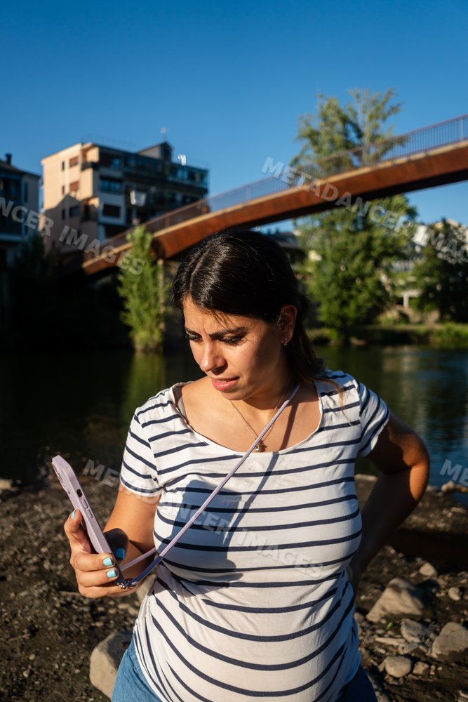 Woman uses phone outdoors by river with bridge and buildings