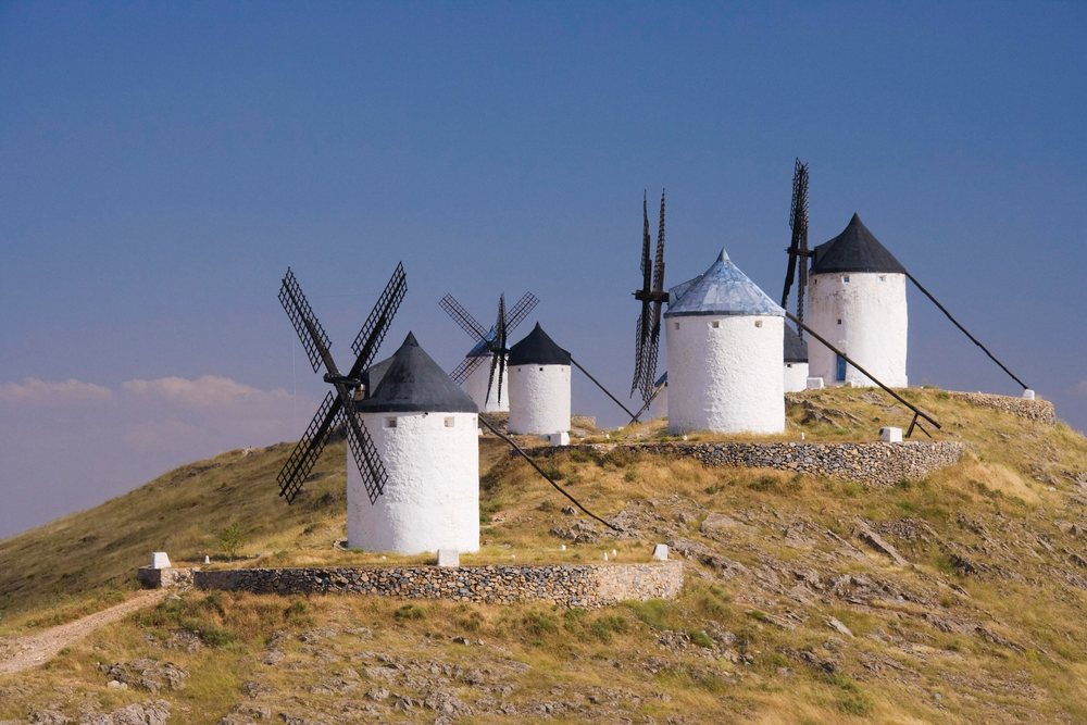 White windmills with conical roofs stand on a rocky hill with stone walls under a clear blue sky, Castilla la Mancha, Spain.