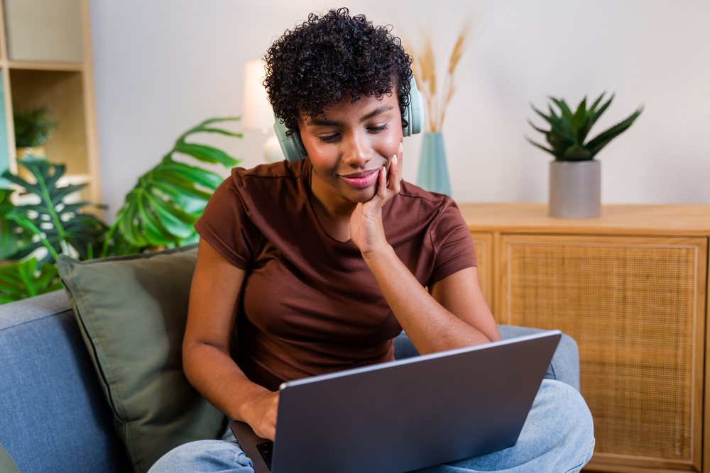 Woman with curly hair wearing headphones uses a laptop while sitting relaxed on a couch indoors at home, Spain.
