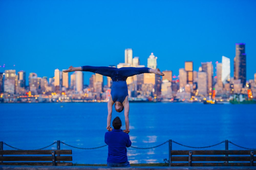 Two people performing partner acrobatics with a handstand supported against the Seattle waterfront cityscape in evening light, USA.