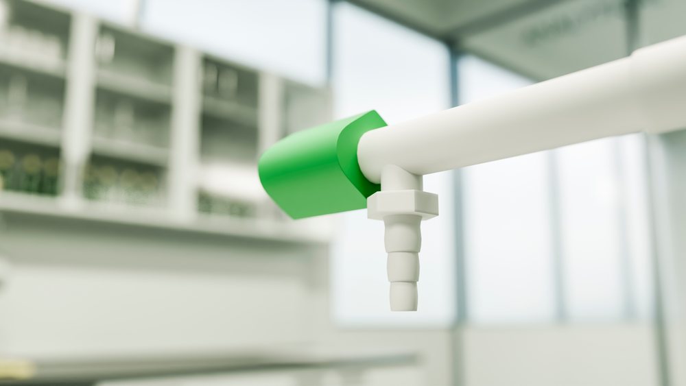 Closeup of a green valve attached to a white pipe fixture in a laboratory setting with a blurry background.