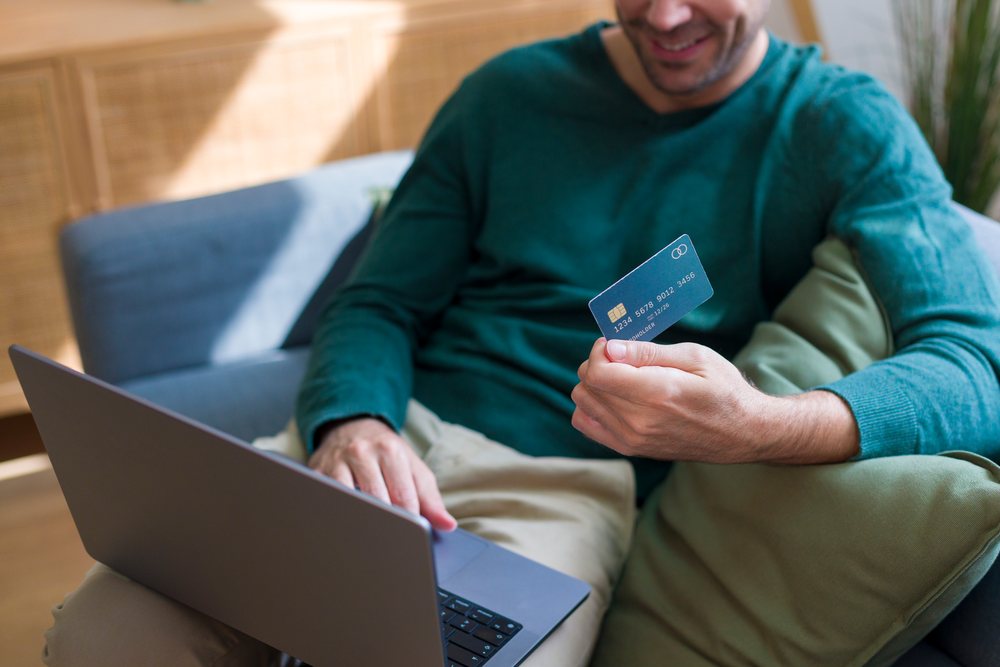 Man sitting on a couch using a laptop and holding a credit card while smiling in a home setting, technology, young adult.