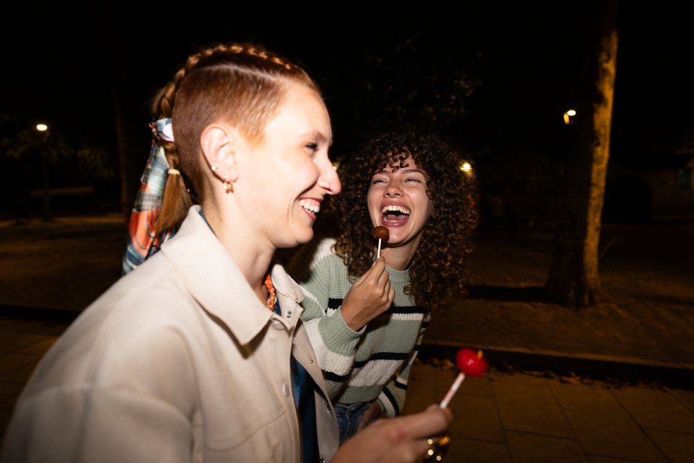 Two women laughing and enjoying lollipops together outdoors at night in a park, friendship, Santiago De Compostela, Spain.