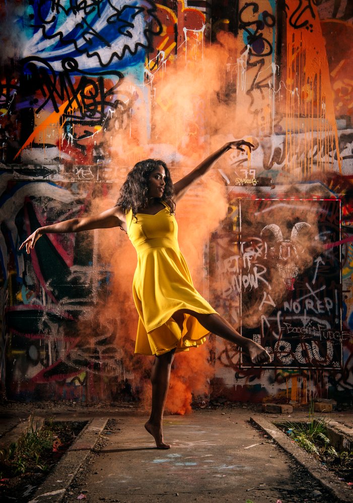 Woman in a yellow dress performs a dance pose with orange smoke in front of a graffiti-covered urban wall, USA.