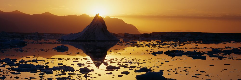 Iceberg floats on calm water reflecting itself and mountainous arctic landscape at sunset under an orange sky.