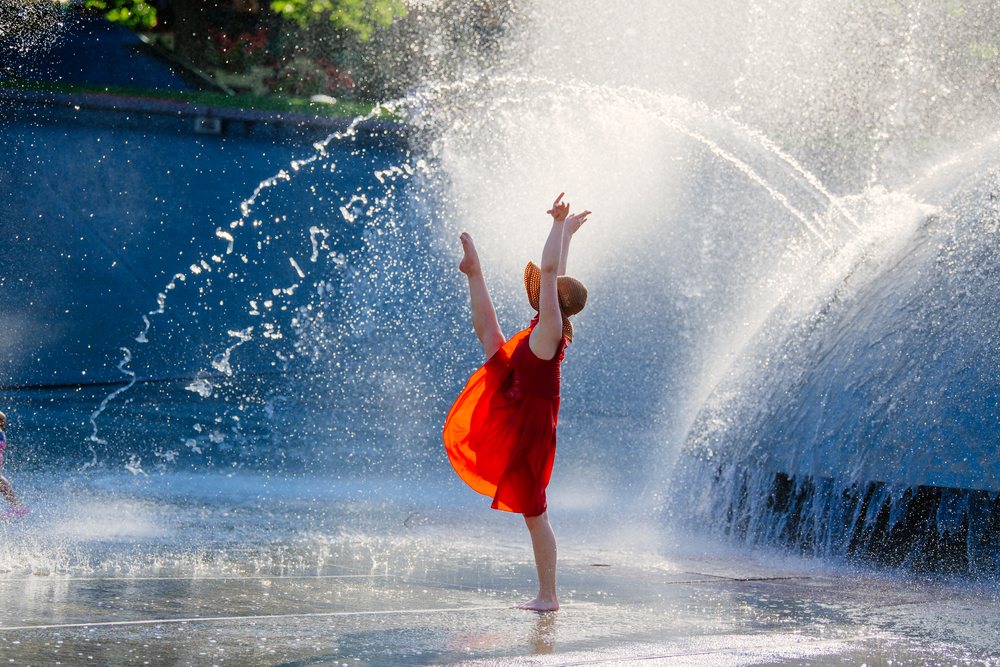 Girl dancing joyfully barefoot in a red dress and sun hat in the water spray of an outdoor fountain on a sunny summer day, USA.