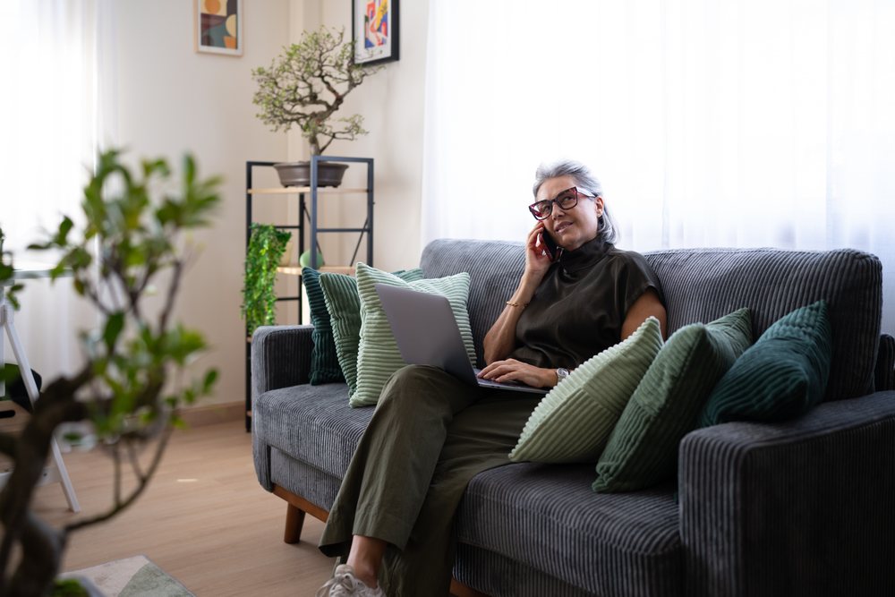 Woman with gray hair wearing glasses sits on a gray couch with green pillows, talking on a phone and using a laptop in a modern Spain living room.
