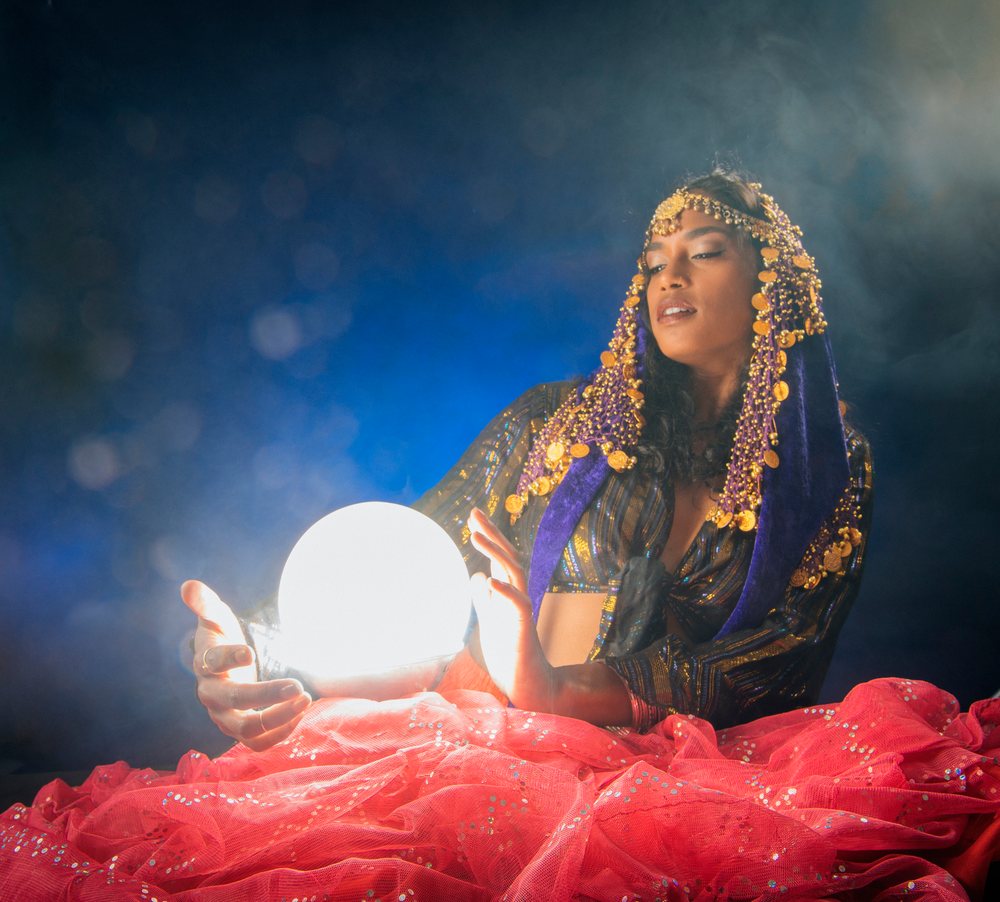 A woman dressed as a fortune teller holds a glowing crystal ball in a mystical indoor setting in the USA with smoke and colorful fabric.
