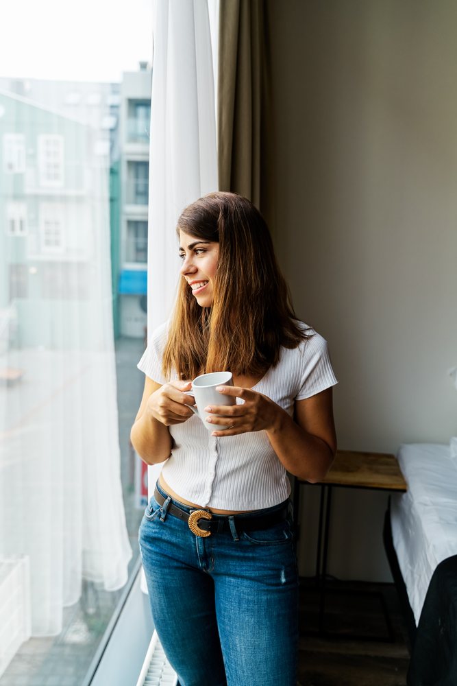 A woman holding a cup looks outside in a hotel room in a hotel