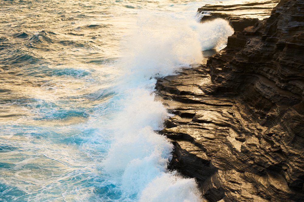 Waves crash against a rocky coastline with sunlight reflecting on the water, Oahu, Hawaii, USA.