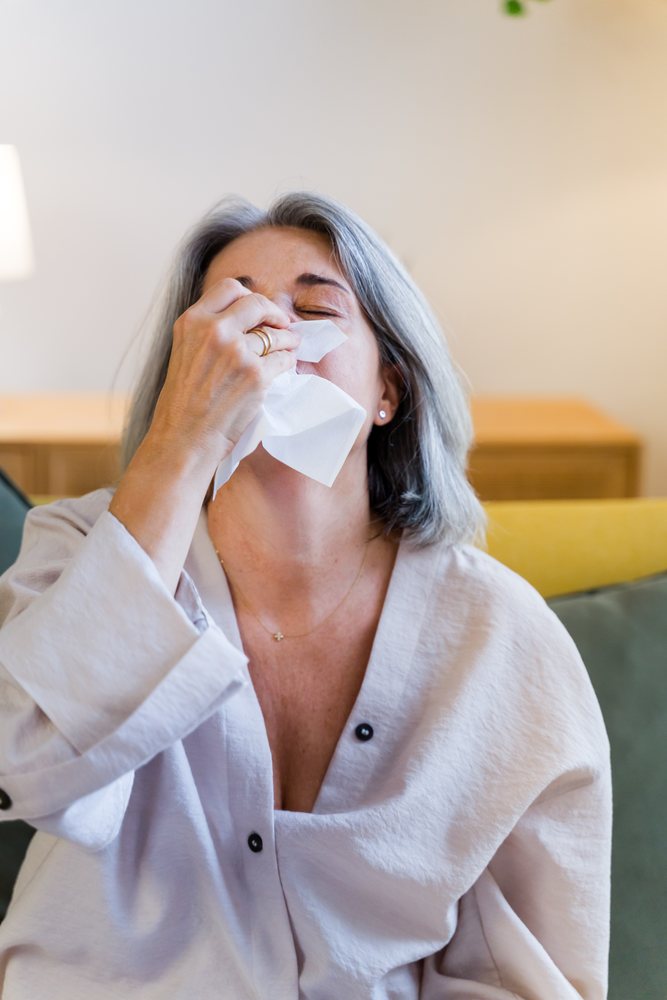Woman with gray hair blows her nose using a tissue while sitting indoors in a living room, Spain.