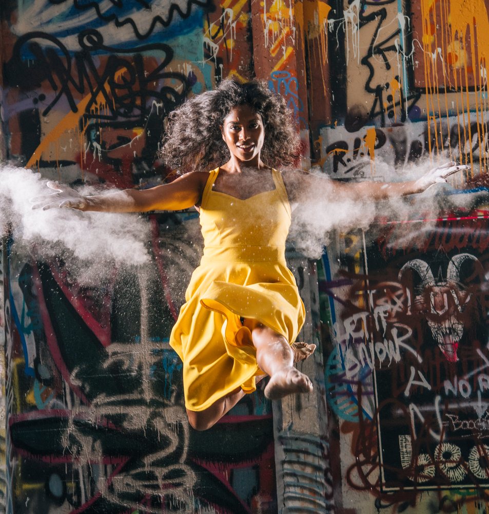 Woman in yellow dress jumps midair amid powder bursts by an urban graffiti wall, USA.
