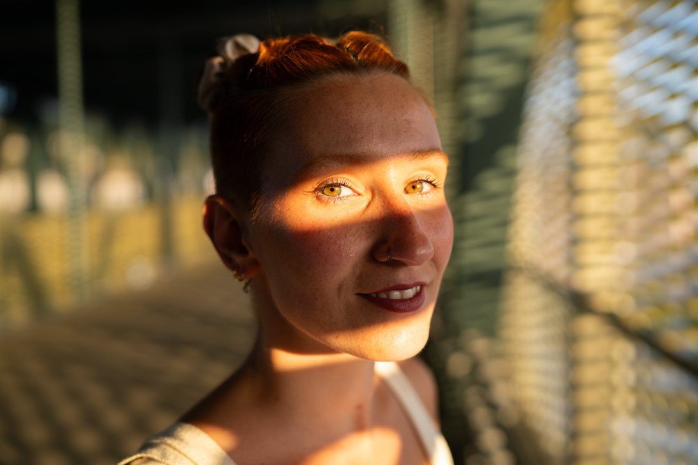 A young woman with red hair smiles as sunlight and shadows from a metal fence create a pattern on her face in an urban setting, Santiago De Compostela, Spain.