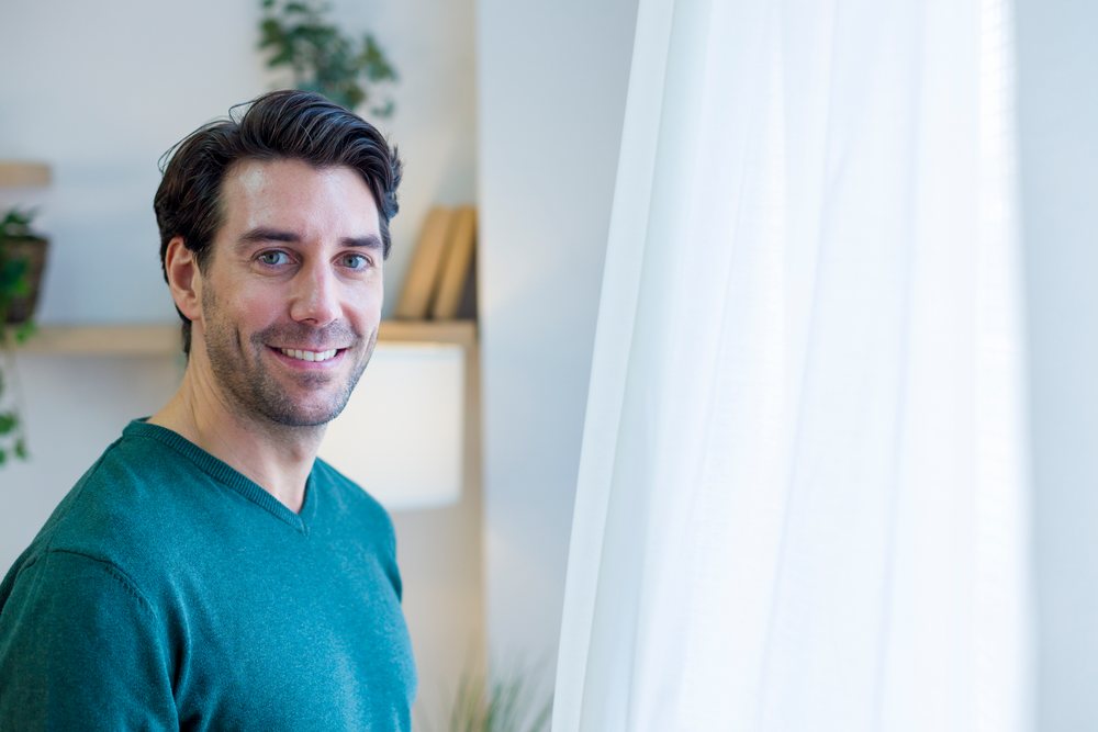 Man smiling and standing indoors near a window with white curtains in a modern home interior, Technology, young adult.