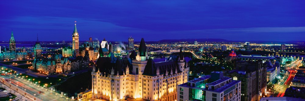 Panoramic night cityscape of Ottawa featuring illuminated Parliament buildings along the river with visible bridge and urban skyline.