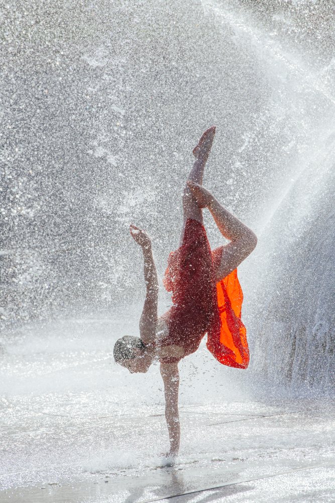 Female dancer performs a one-handed handstand through water spray at an outdoor fountain, USA.