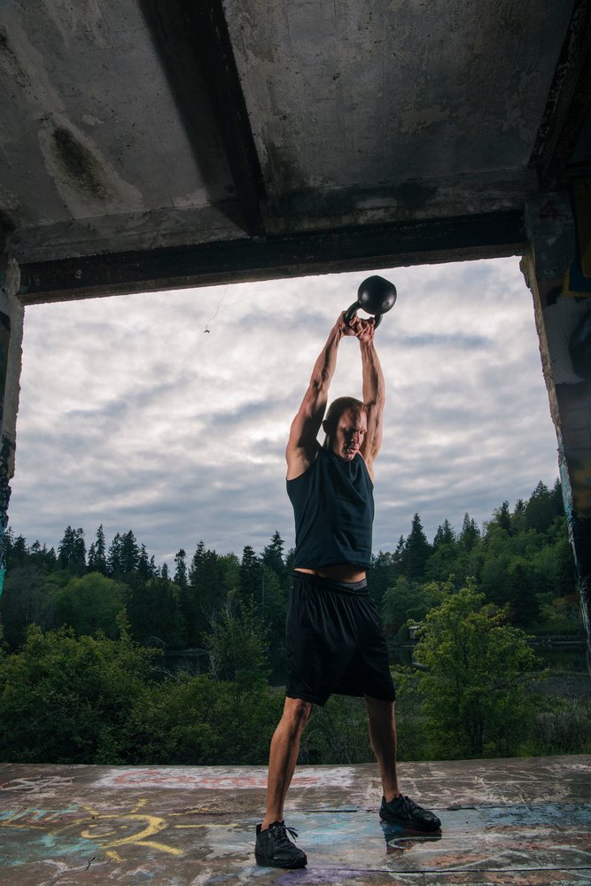 Man holding a black kettlebell overhead during an outdoor workout inside an abandoned graffiti-covered structure, USA.