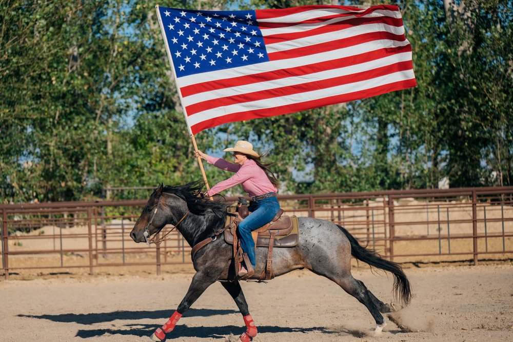 Cowgirl gallops on a horse waving a large American flag in an outdoor riding arena, Southwest USA.