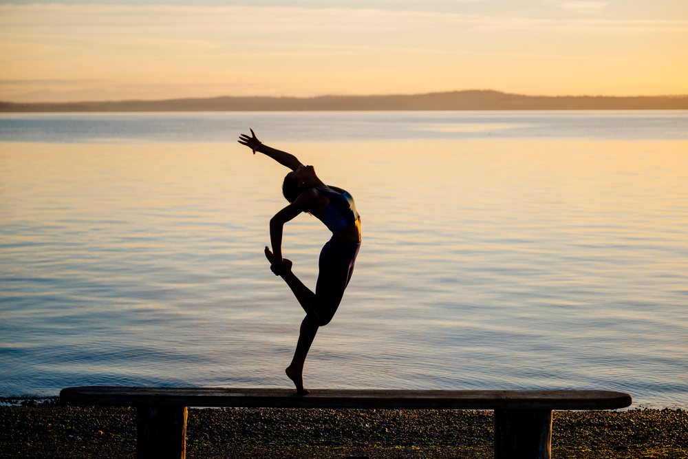 Woman practicing yoga acrobatics stretching pose on a bench by the water at sunset, USA.