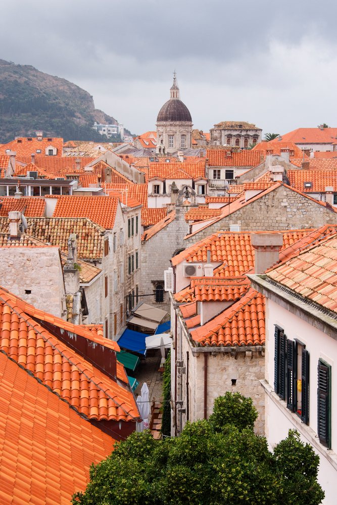 View of old city rooftops with red tiles, historic stone buildings, a church dome, and green foliage under a cloudy sky in Dubrovnik, Croatia.
