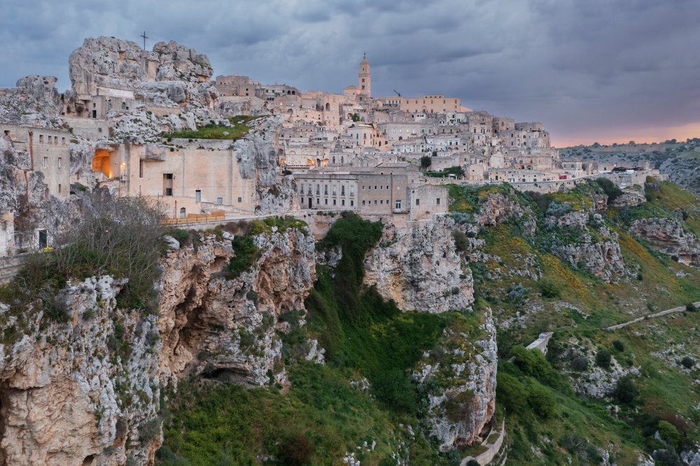 Historic stone buildings of Sasso Caveoso cluster on rocky cliffs with the Church of San Pietro Caveoso visible at Dawn under a cloudy sky, Matera, Basilicata, Italy.