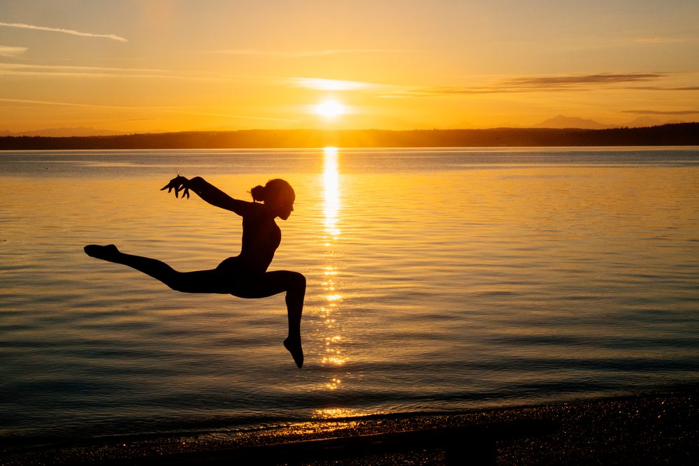 Silhouette of a dancer leaping by the water during a vibrant orange sunset, yoga, acrobatics, USA.