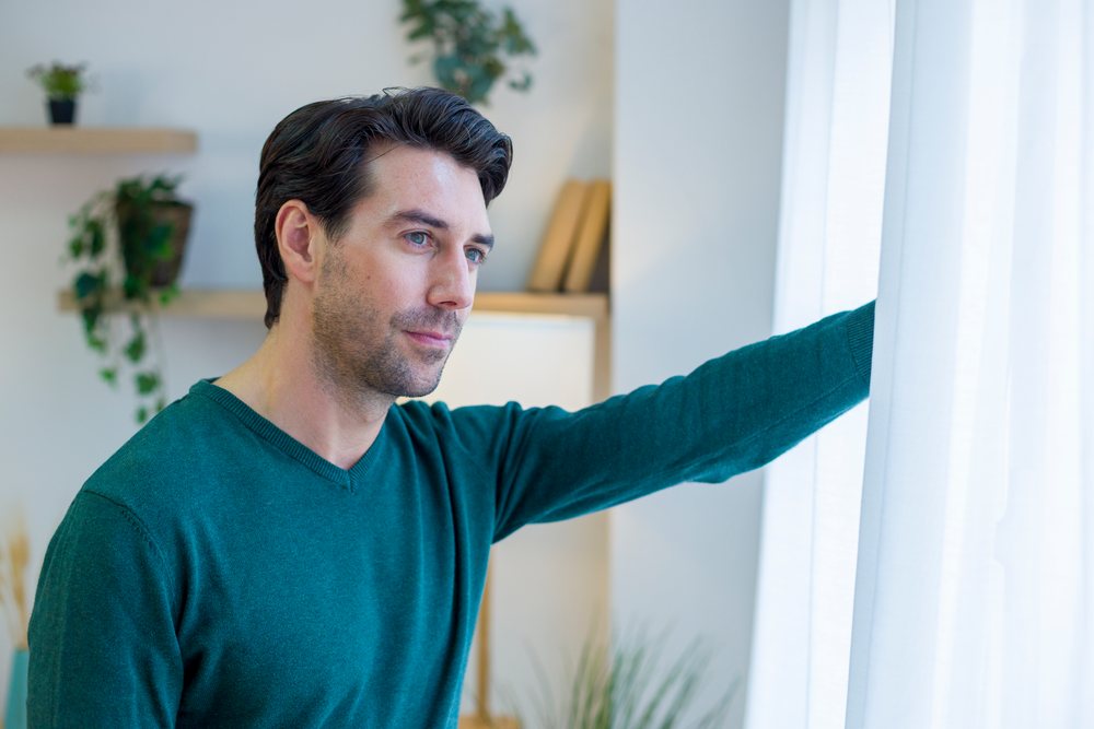 Man wearing a teal sweater stands indoors looking thoughtfully out a window, holding a white curtain in a living room with plants and books on a shelf, technology, young adult, home.