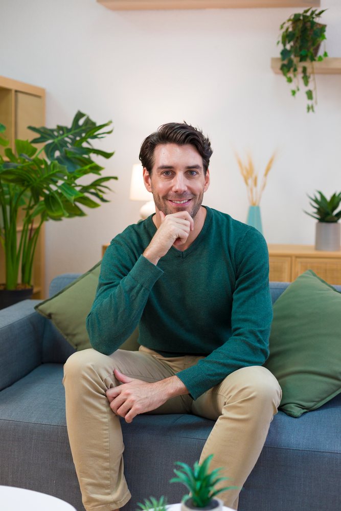 Man wearing a green sweater and beige pants sits comfortably on a gray sofa with green cushions in a modern living room with plants and wooden furniture, technology, young adult, home.
