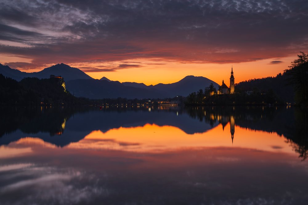 Church of the Assumption of the Virgin Mary and Bled Castle silhouetted at Sunrise over Lake with reflected mountains and sky, landscape, cityscape, Slovenia.