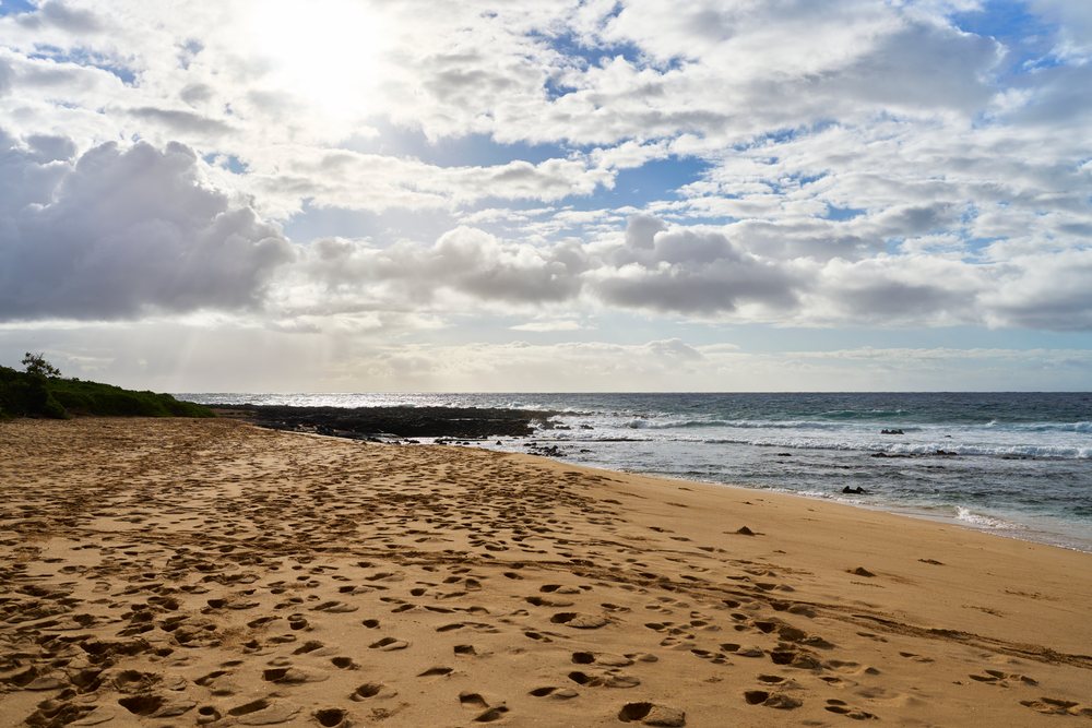 Beach with sandy footprints beside ocean waves under a partly cloudy sky, Hawaii, USA.
