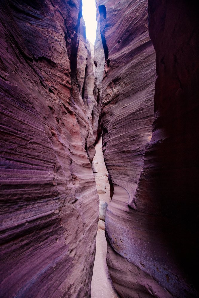 Narrow slot canyon with layered rock walls and a sandy floor under sunlight filters through the top opening, Buckskin Gulch, Utah, USA.
