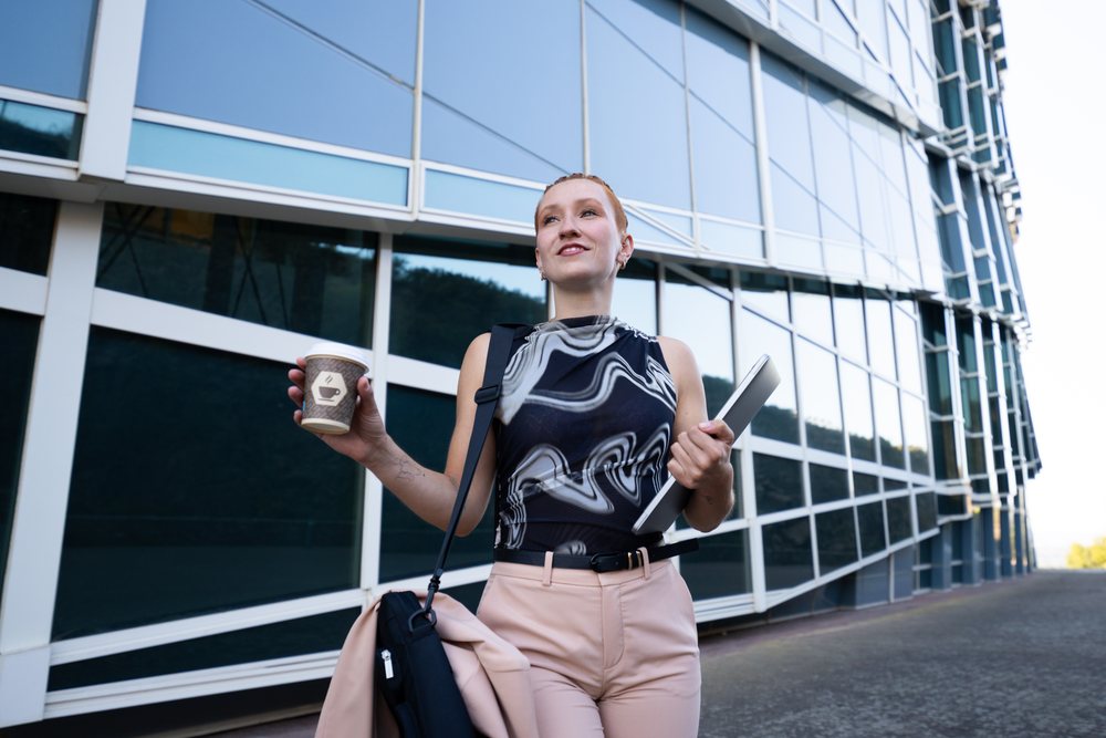 Businesswoman walking outdoors near a modern glass office building holding a coffee cup, laptop, and carrying a bag, businesswomen, Spain.