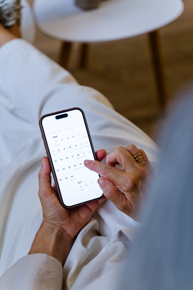 Person holding smartphone displaying a calendar app indoors wearing white pants, sitting near a table with a brown floor visible, Spain.