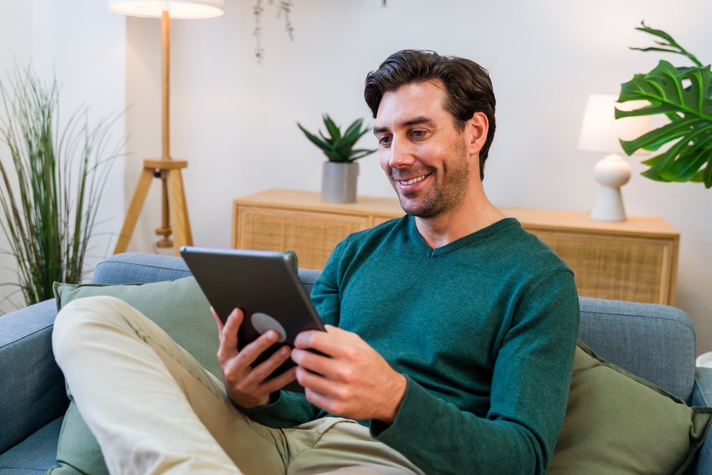 Man sitting on couch in a modern indoor living room holding a tablet and smiling, Spain.