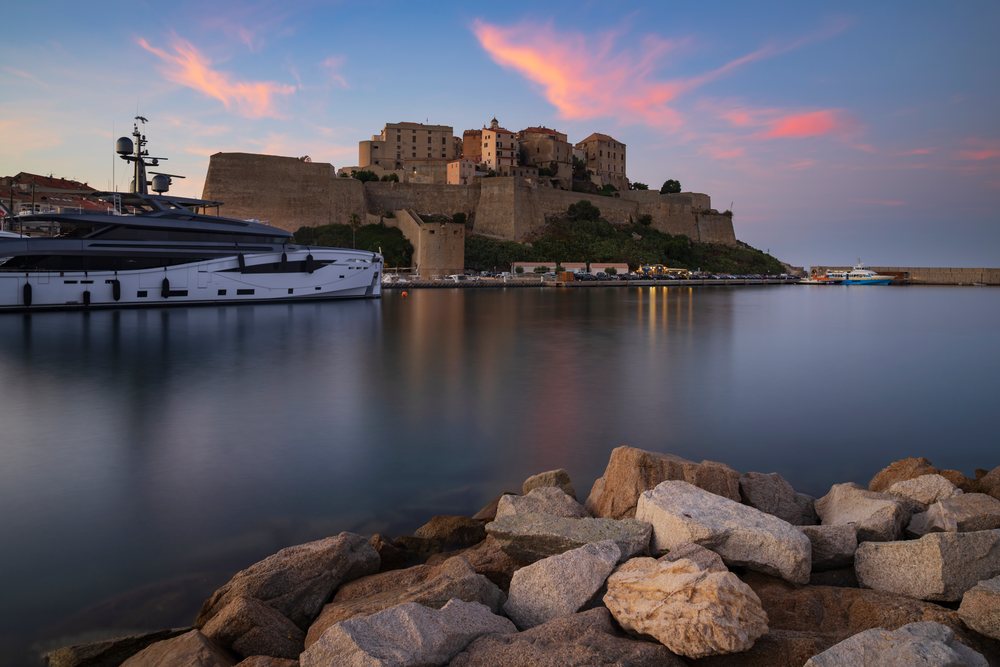 Citadel overlooking harbor at Sunset from yacht moored at mole with calm water and rocky shore, Travel Photography, Calvi, Corsica, France.