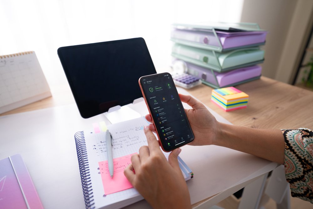 Person using smartphone at home office desk with tablet and notebook, Spain.