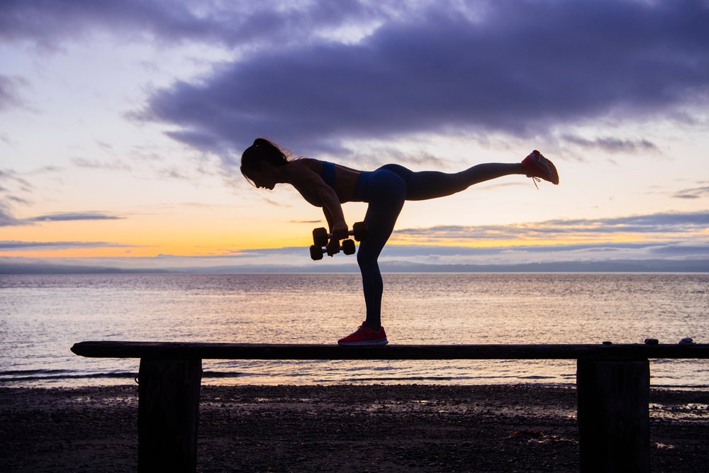 Woman balances on one leg on a bench while holding dumbbells during outdoor exercise at sunset on a beach, WA, USA.