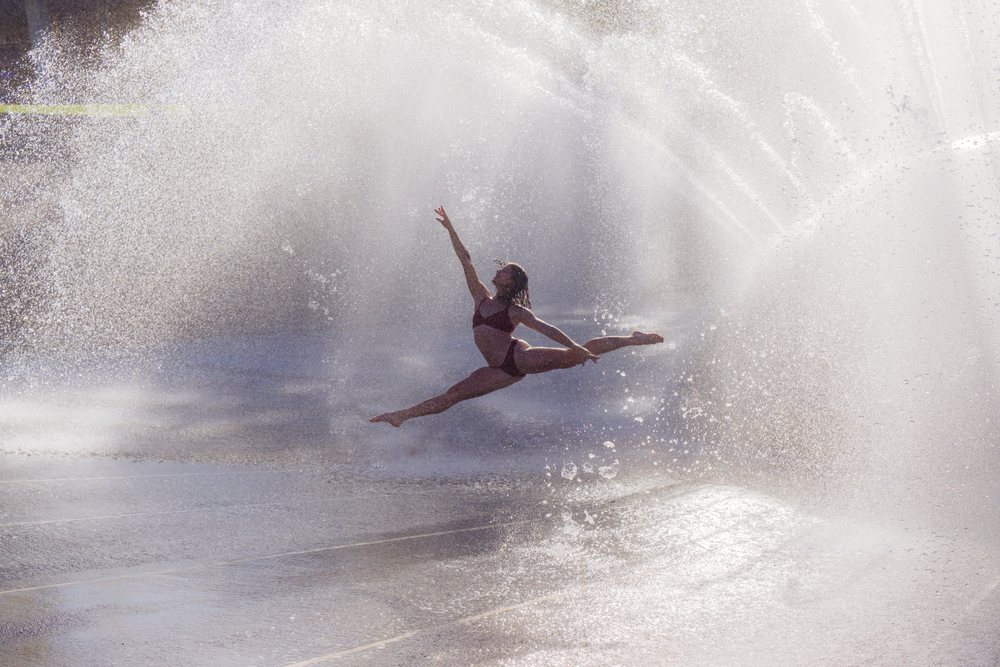 Woman in swimsuit performing a split leap through water spray from a fountain outdoors, partner, acrobatics, yoga, USA.
