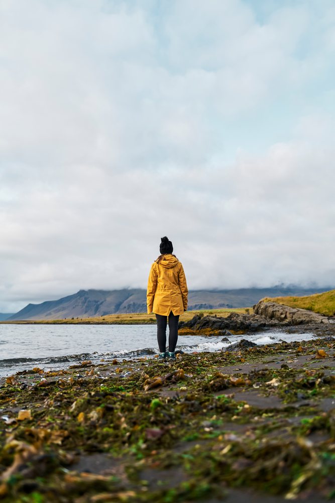 A white woman in a yellow jacket stands on a rocky shore