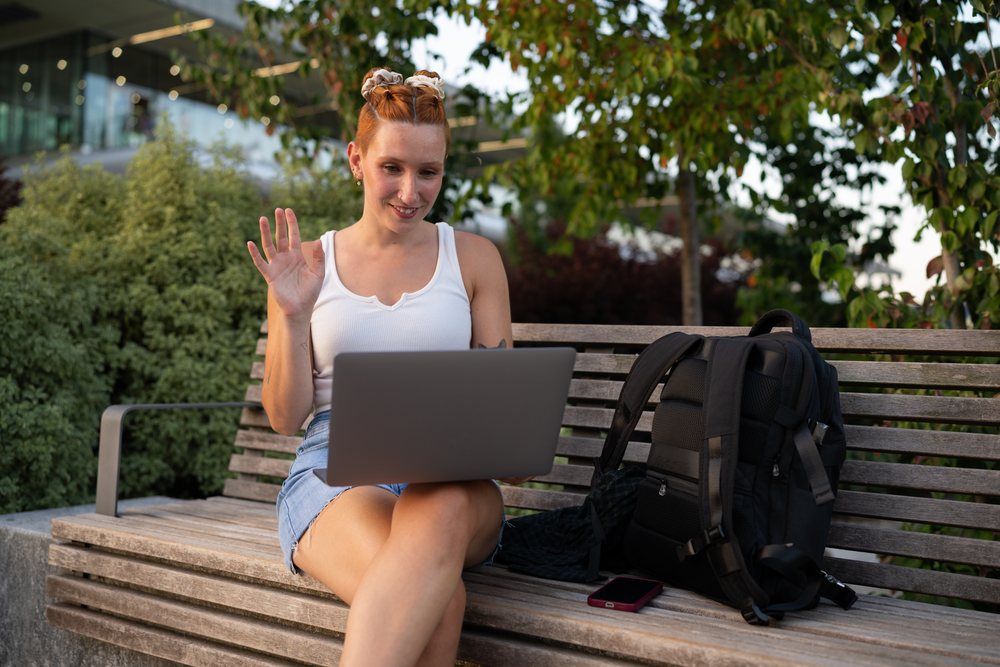 A young woman wearing casual summer clothes waves during a video call on her laptop while sitting on a bench in an urban park area, Santiago De Compostela, Spain.