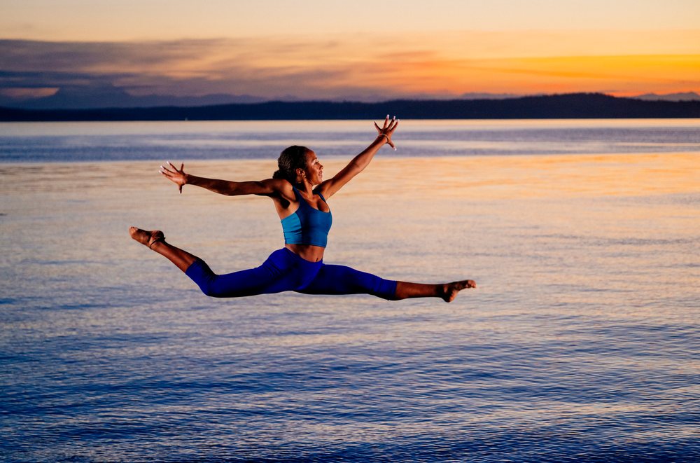 Woman in blue athletic wear performing a split jump over calm water at sunset, yoga, acrobatics, USA.