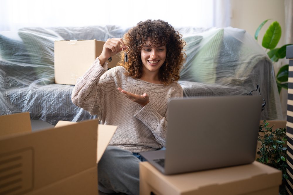 Smiling woman sitting on floor holding keys near laptop and cardboard boxes during home move, moving, Spain.
