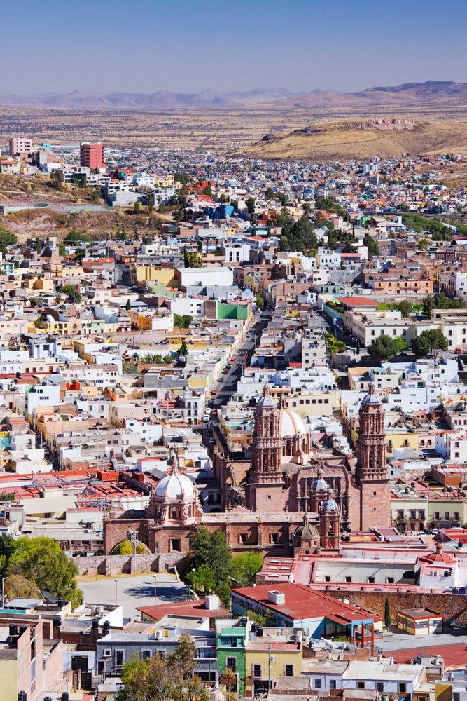 Clear daytime cityscape of Zacatecas featuring a historic church with multiple domes and towers amid urban buildings and desert mountains, Mexico.