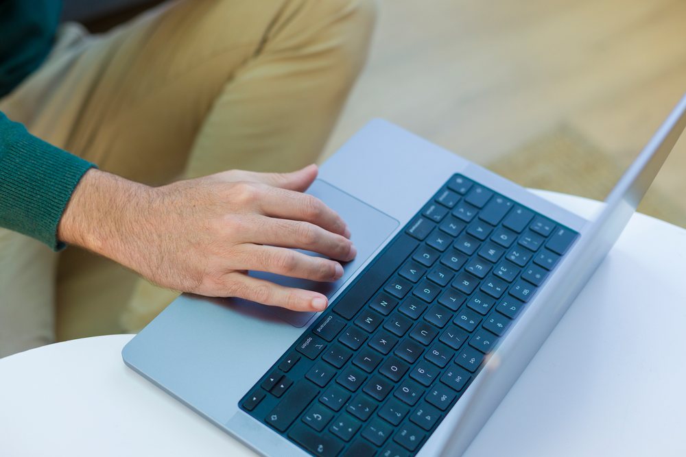 Young adult person uses a laptop and trackpad indoors at home, wearing beige pants and a greenish-blue top.