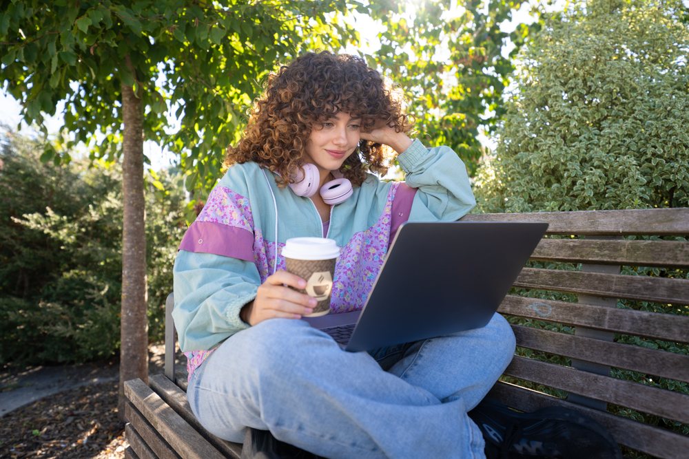 Woman sitting on a wooden bench in a park in Spain using a laptop and holding a coffee cup.