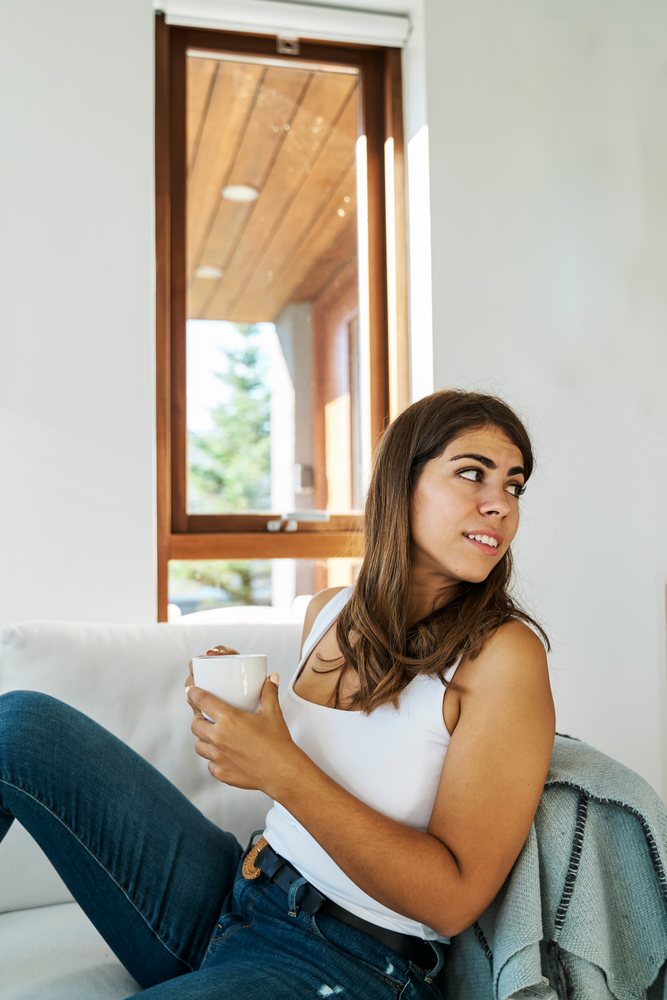 A young white woman relaxes on a couch holding a mug by a window